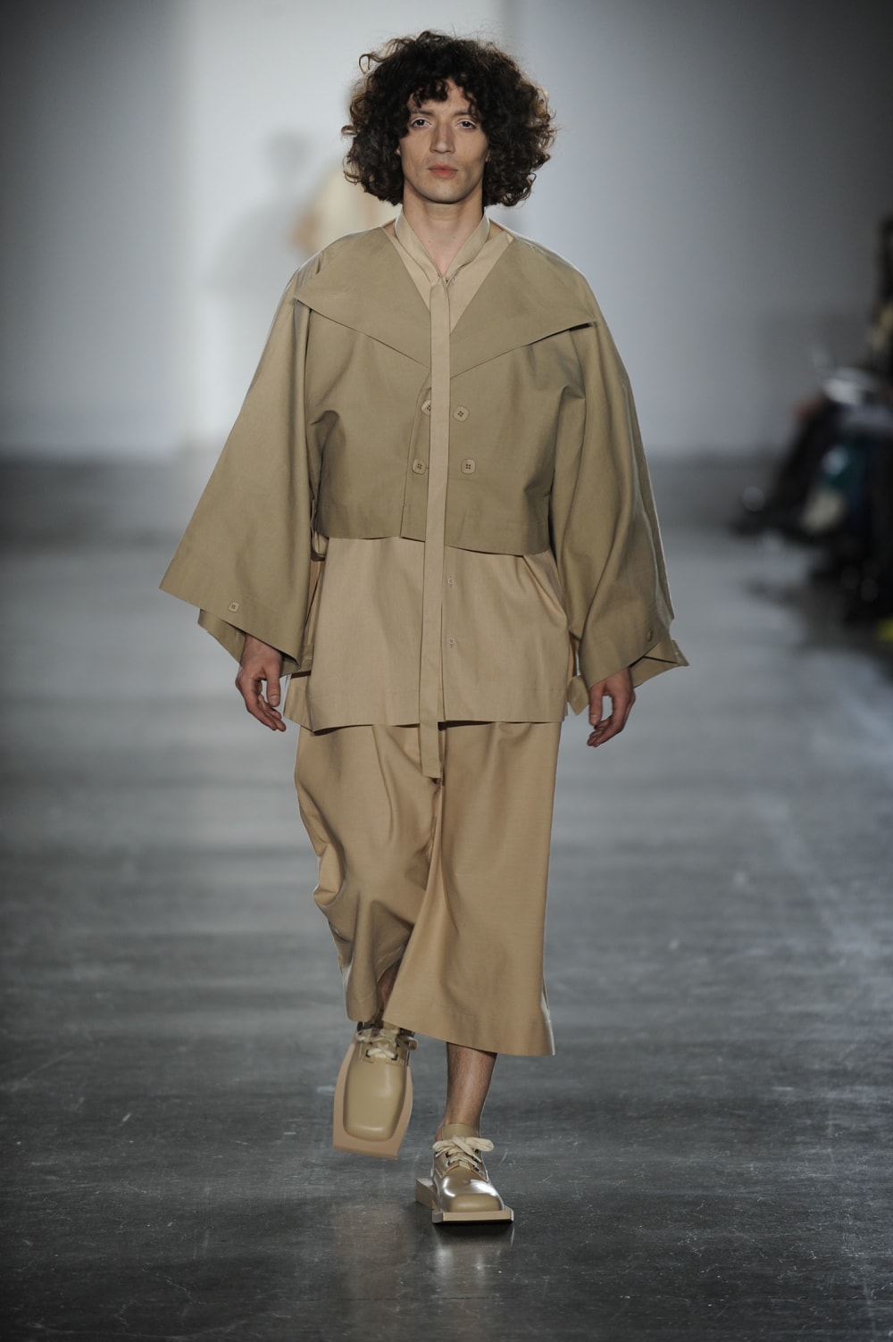 Man on runway wearing different shades of beige with baggy cropped bell sleeved jacket with 2 double breasted buttons and button detailing on sleeves, long shirt top with buttons, long thin tie, baggy long shorts and squared off laced shoes 