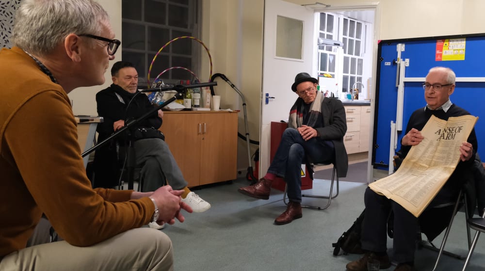 An interview taking place. A man is holding up a newspaper, and the 3 other people are listening to him talk. 