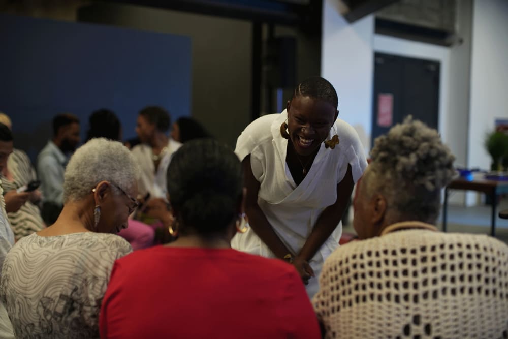 Image shows a woman bending down and laughing while speaking to a group of women who are sitting down