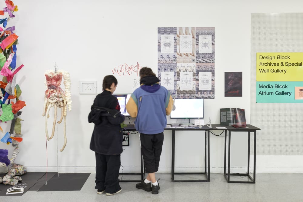 Two people stand at a display table in an art gallery, viewing digital screens and mixed-media artworks on a white wall decorated with posters, signs and a sculptural piece resembling a human torso.