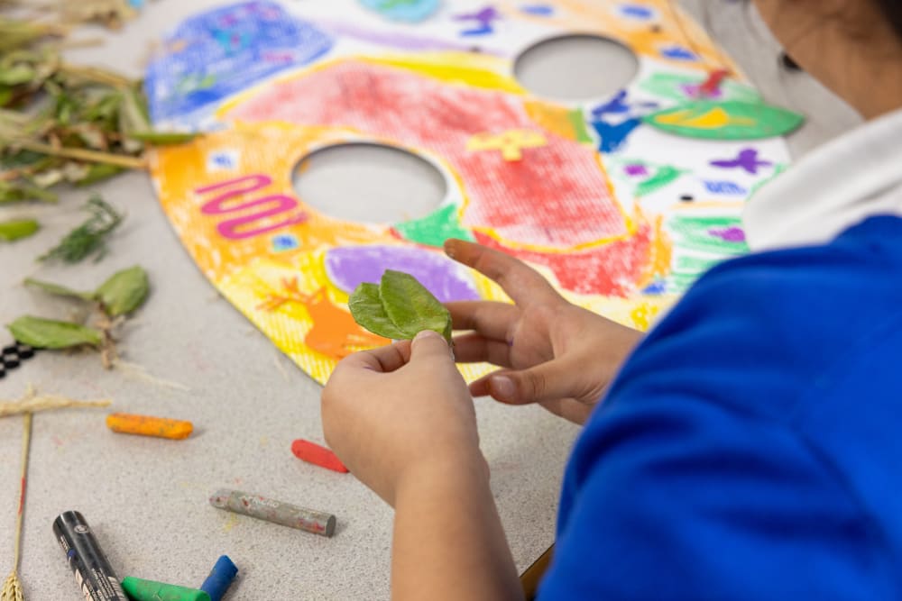 A child holding 2 leaves in their hand sticking it to a cardboard cut out of an apple coloured in with crayons.