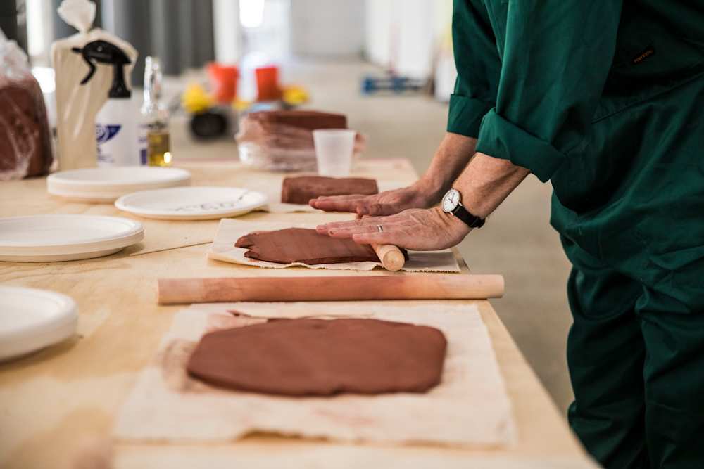 A side shot of a person in a green boilersuit rolling clay on a worktop.