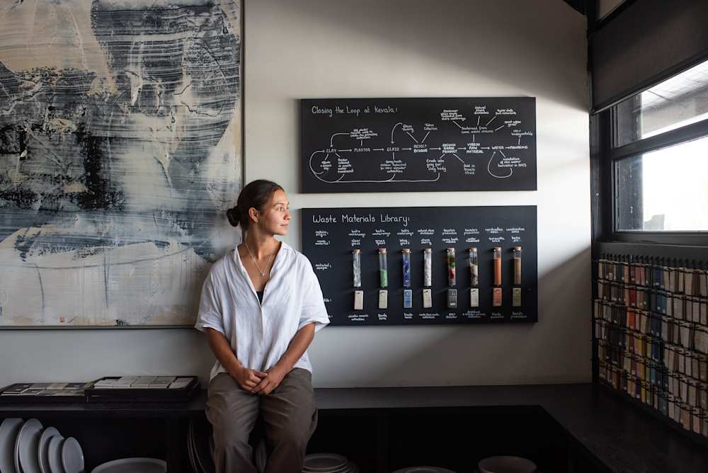 A person sits on a bench in a ceramics studio, looking toward a window, beside wall‑mounted display boards showing a “closing the loop” process diagram and a “waste materials library” with labelled samples in glass tubes; shelves of material swatches and ceramic plates are visible around the space.