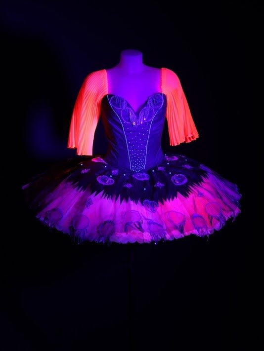 Photo of a tutu glowing in a black room. The tutu has a purple bodice and skirt, and pink/orange arms. 