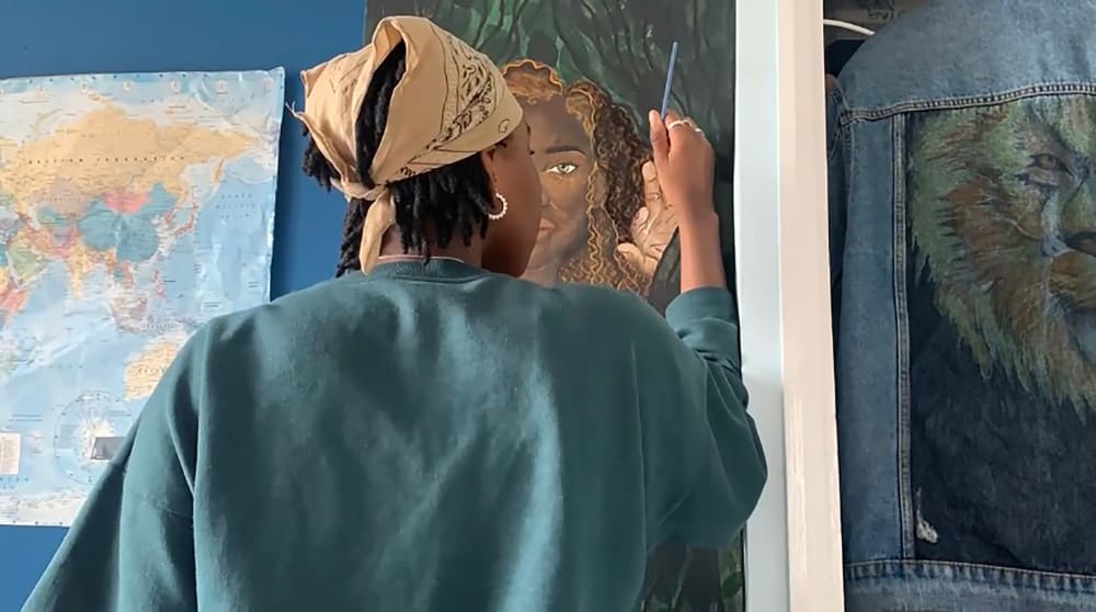 Photograph of the back of Emanuela, a black woman wearing a beige headscarf and green jumper. She is holding a paintbrush, painting a portrait of her studio.