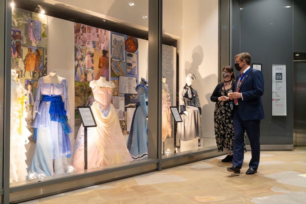 A man in a suit and a young woman in a flora dress, both in face masks, stand in front of a large shop window through which five mannequins wearing elaborate costumes are visible.