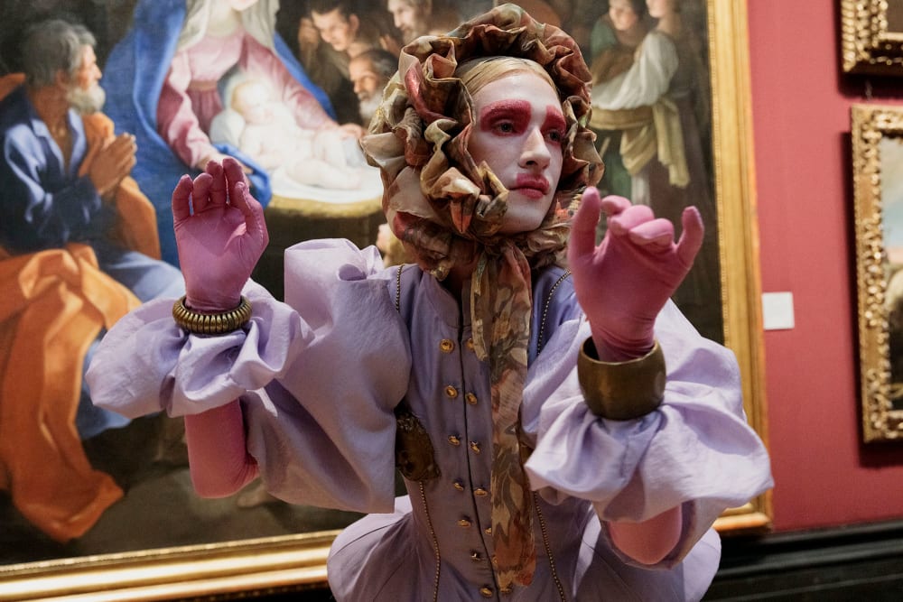 A person in pink make-up wears a Baroque-inspired costume in front of a large painting.