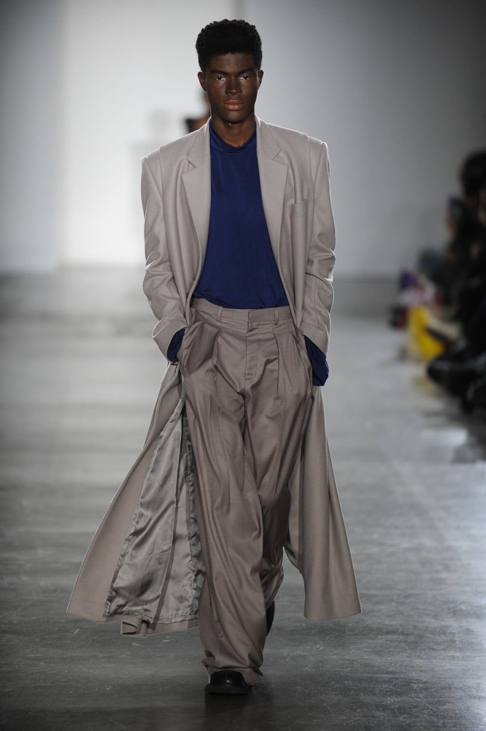 Man on runway wearing long warm grey smart jacket and matching straight baggy fit pants with blue full sleeved top and black shoes