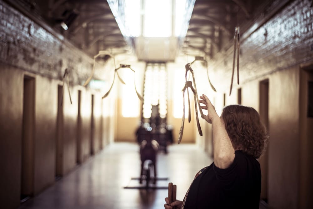 The image shows Janaleen Wolfe in the foreground, mobility cane held in her right hand, her left hand reaching up to ‘ring’ one section of Aaron McPeake’s Resonant Cuts (2011), a series of suspended bell slices, cast in bell bronze. The seven slices were installed by Fayen D’Evie in one of the imposing top-lit corridors of the nineteenth century Castlemaine Gaol, as part of her collaborative project From Dust to Dust. Behind Janaleen, we see the space with a series of repetitive doors to cells, and a blurred staircase leading up to a mezzanine level of the former prison.