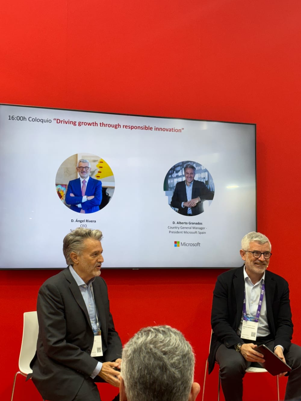 Two people seated in front of an audience in conversation, in front of a red wall. 