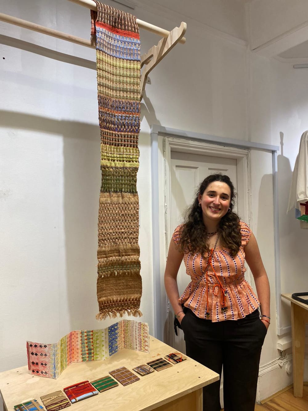 Image shows a person standing in a gallery next to a table of textile work and a hanging woven piece of fabric.