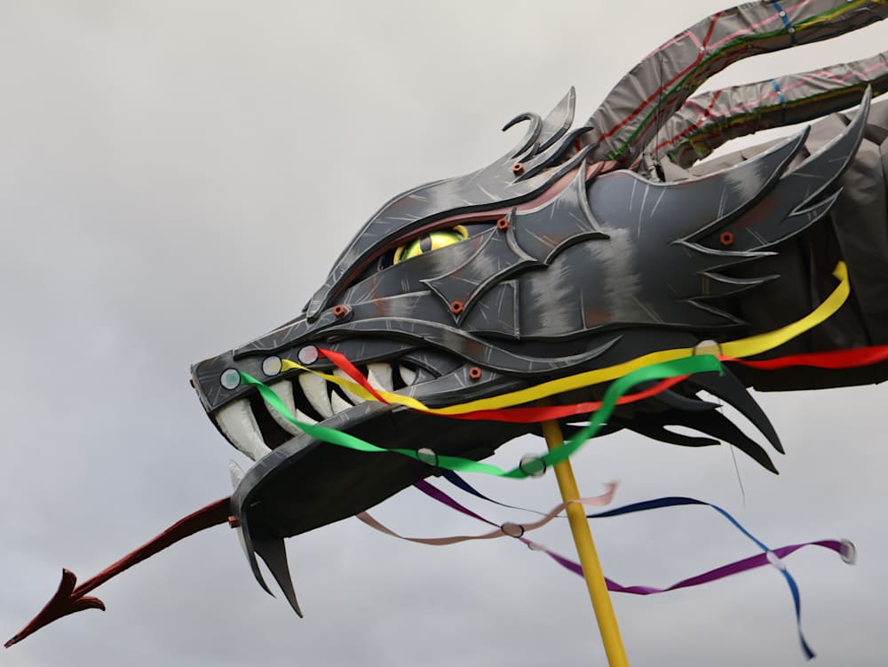 Alt text: A colorful dragon kite with a fierce expression soars against a gray sky. Its sharp features and flowing ribbons create a dynamic, lively scene.