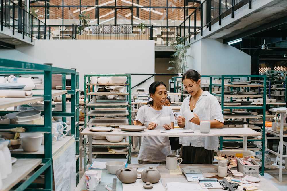 Two people stand at a worktable in a ceramics studio, examining a white cup, surrounded by shelves of drying ceramic plates and bowls; tools, teapots, and paperwork are spread across the table, with a large open workshop space and mezzanine level in the background.