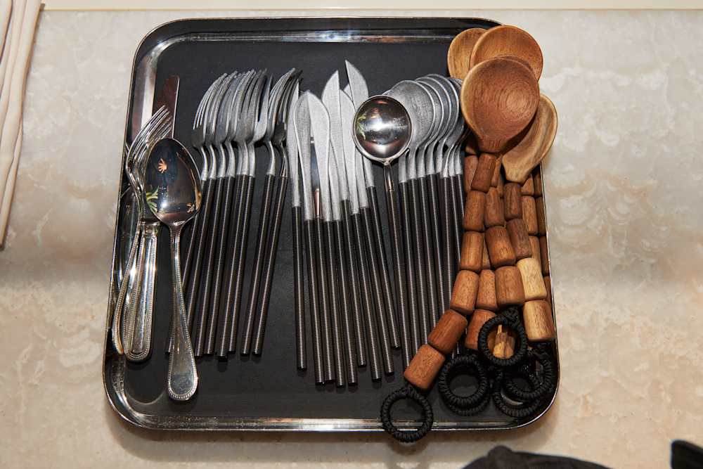 A neatly arranged tray with shiny metal cutlery and round wooden spoons on a marbled surface. The tone is organized and functional.