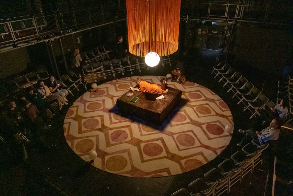 Image shows a birds eye view of the set for 'The Father'. From this view, a circular 1970s inspired rug can be seen with a large circular orange fringed lampshade hanging over brown square seating. An actor can be seen lying beneath the light on the seating and audience members can be seen taking their seats in the background.