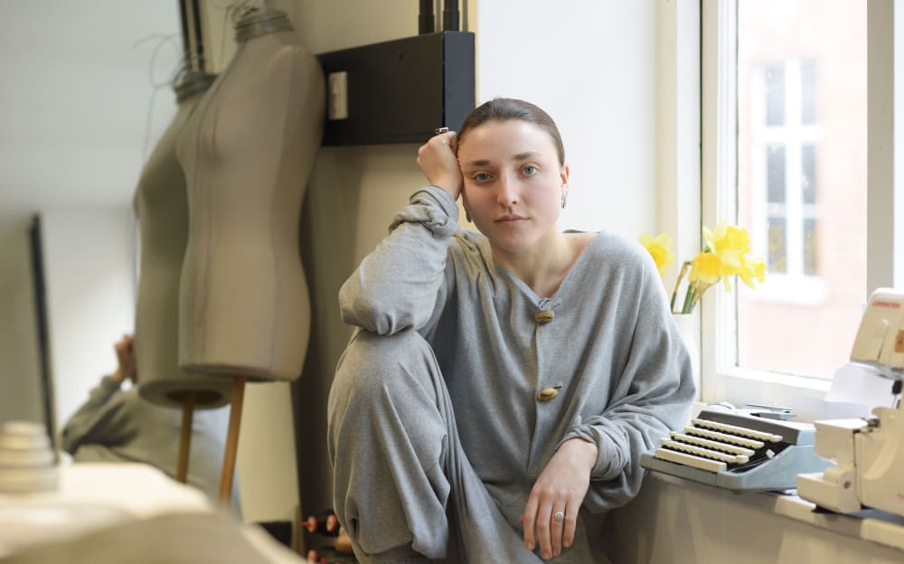 Young person in gray loungewear sits by a window with daffodils, resting their head on a hand. A mannequin and typewriter are nearby, creating a calm, contemplative atmosphere.