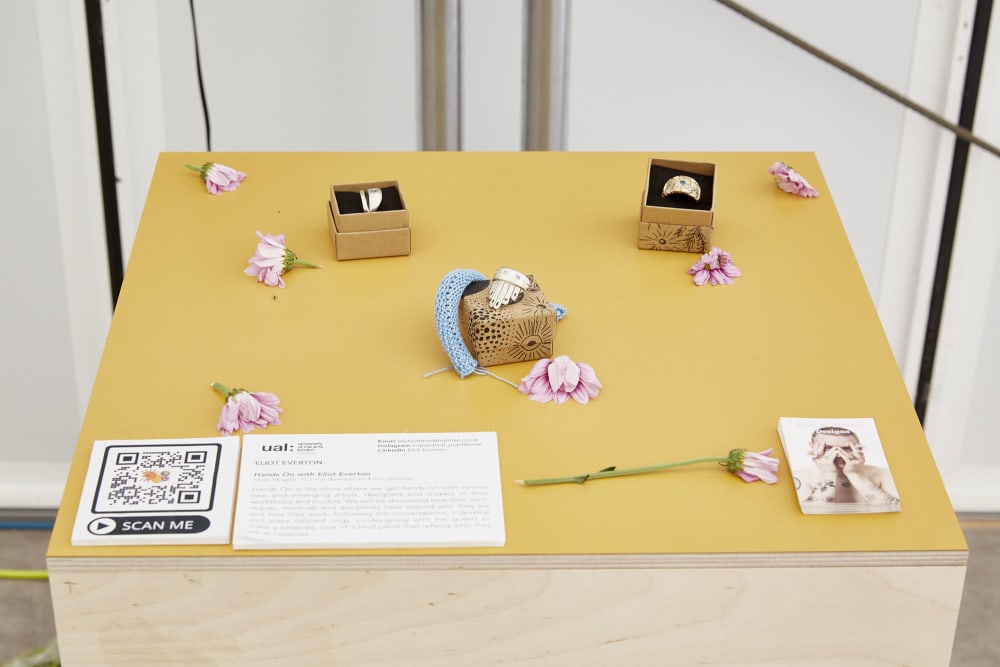 A yellow topped wooden podium displays 3 gold rings designed by Eliot. The rings are placed with their ring boxes and are surrounded by pink flowers. In the bottom right corner, a pile of business cards displaying photos of Eliot are positioned, and to the left corner a QR code with the words SCAN ME is displayed alongside some text on a white card. 