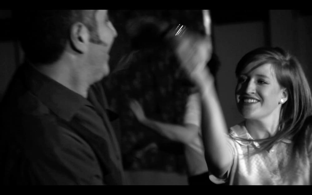 Black and white still of a woman smiling and moving her arms. To her left is a man