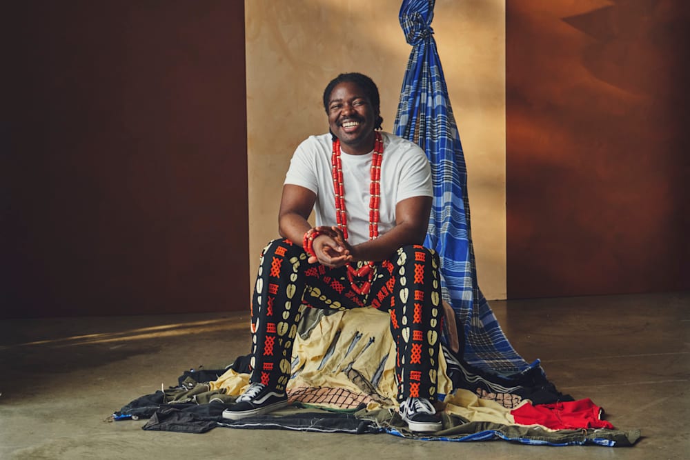 Person seated on layered textiles wearing patterned trousers and beaded jewellery, with draped fabric in the background.