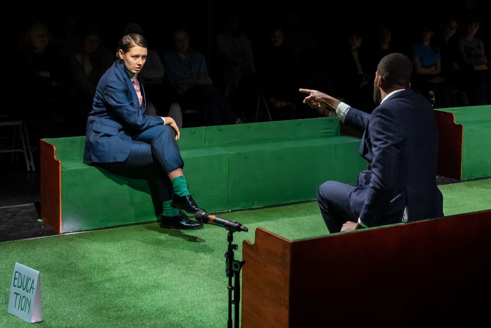Image shows 2 people in a set designed to look like the United Kingdom's House of Commons. It is green with benches on either side. 1 person can be seen sitting on the bench facing the other person and pointing in an accusatory manner. 