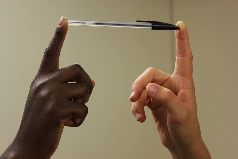 A photo of 2 hands, one black and one white holding a biro pen between the index fingers