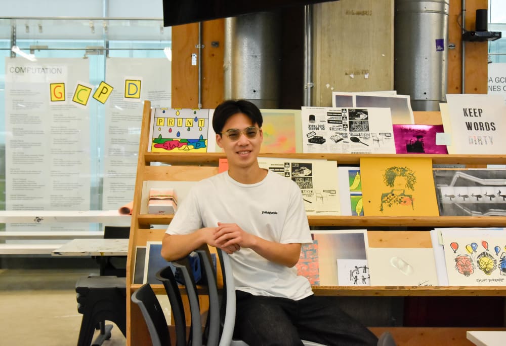  An image of student Tripoom Chongpanisook at the graphic communication studios at CSM. Tripoom is seated on a chair, in front of a rack of magazines.