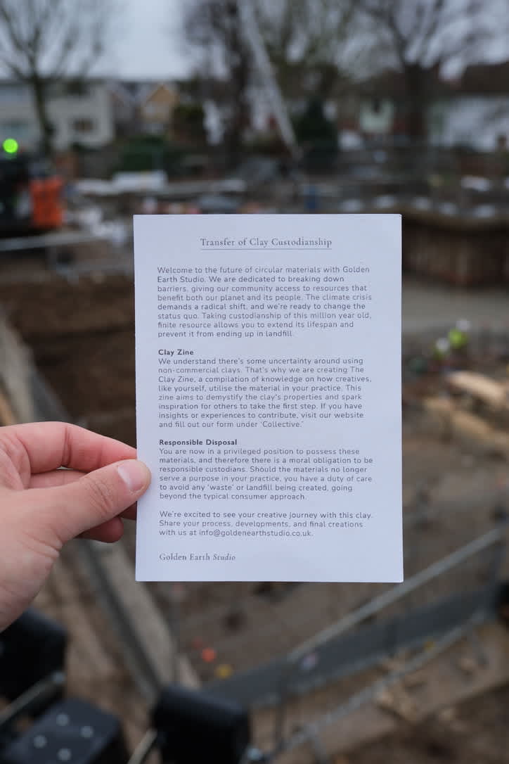 A hand holds a printed card titled “Transfer of Clay Custodianship” in the foreground, with paragraphs of text about circular materials, a clay zine, and responsible disposal, set against a blurred outdoor scene of a community garden or work site with raised beds and tools in the background.