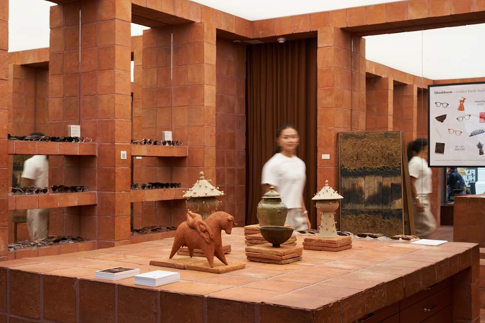 A gallery-like retail interior built from warm terracotta tiles displays ceramic objects and sculptures on a central counter, with shelves of glasses lining the walls and a person walking through the space; framed artwork and a poster reading “bloobloom × Golden Earth Studio” are visible in the background