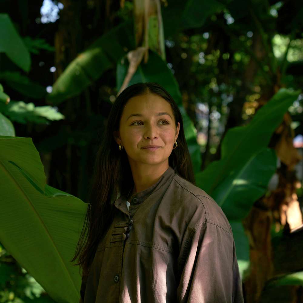 A person stands among large green tropical plants, wearing a loose button-up shirt, with dappled light filtering through the foliage in the background.