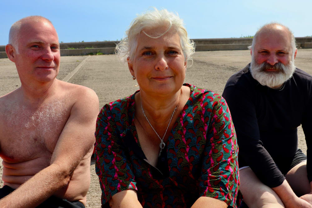 A photograph of 3 people, a man, a woman and another man sitting on a beach 