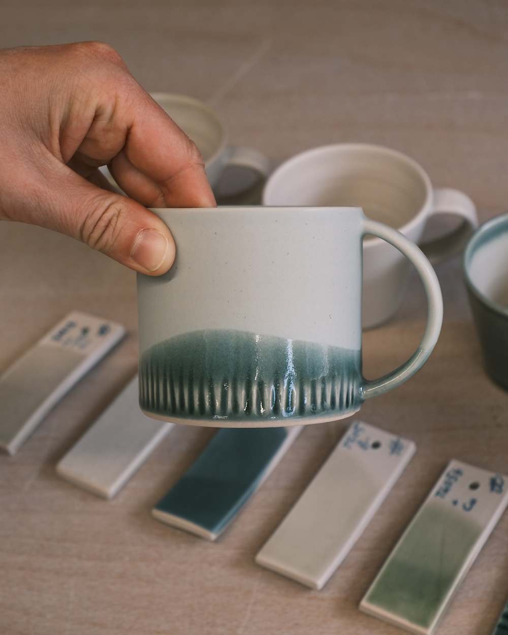 A hand is holding a coffee mug showing a glazed base. More coffee cups and small glazed tiles are beneath.