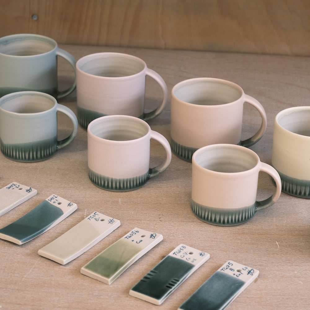 Two rows of coffee mugs with glazed bases. Next to them are glaze labels indicating the different colour glazes of the bases