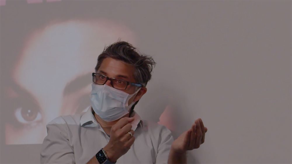 A director wearing a face mask sits in front of a projected screen.