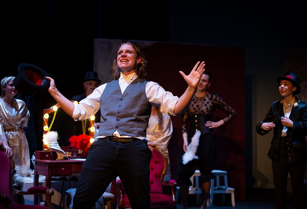 An actor wearing a grey waistcoat and white shirts stands looking out to the audience with his arms raised to the side and a happy expression on their face.