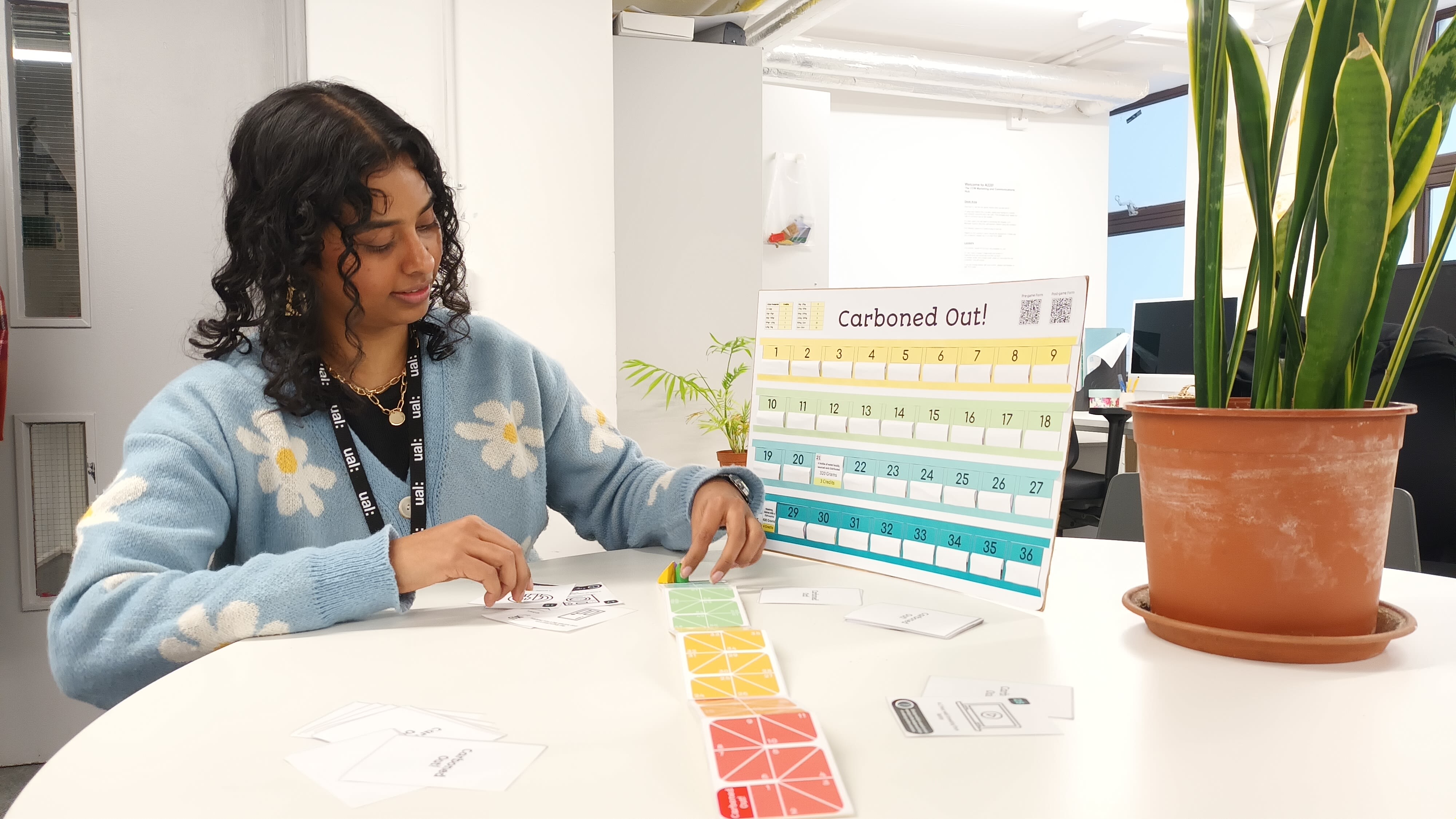 Nanthini sits at a table wearing a blue cardigan with white flowers on. Next to her is a large piece of card which says ‘Carboned Out’. On the table are playing cards and game counters. To left of her is a large plant in a brown plant pot.