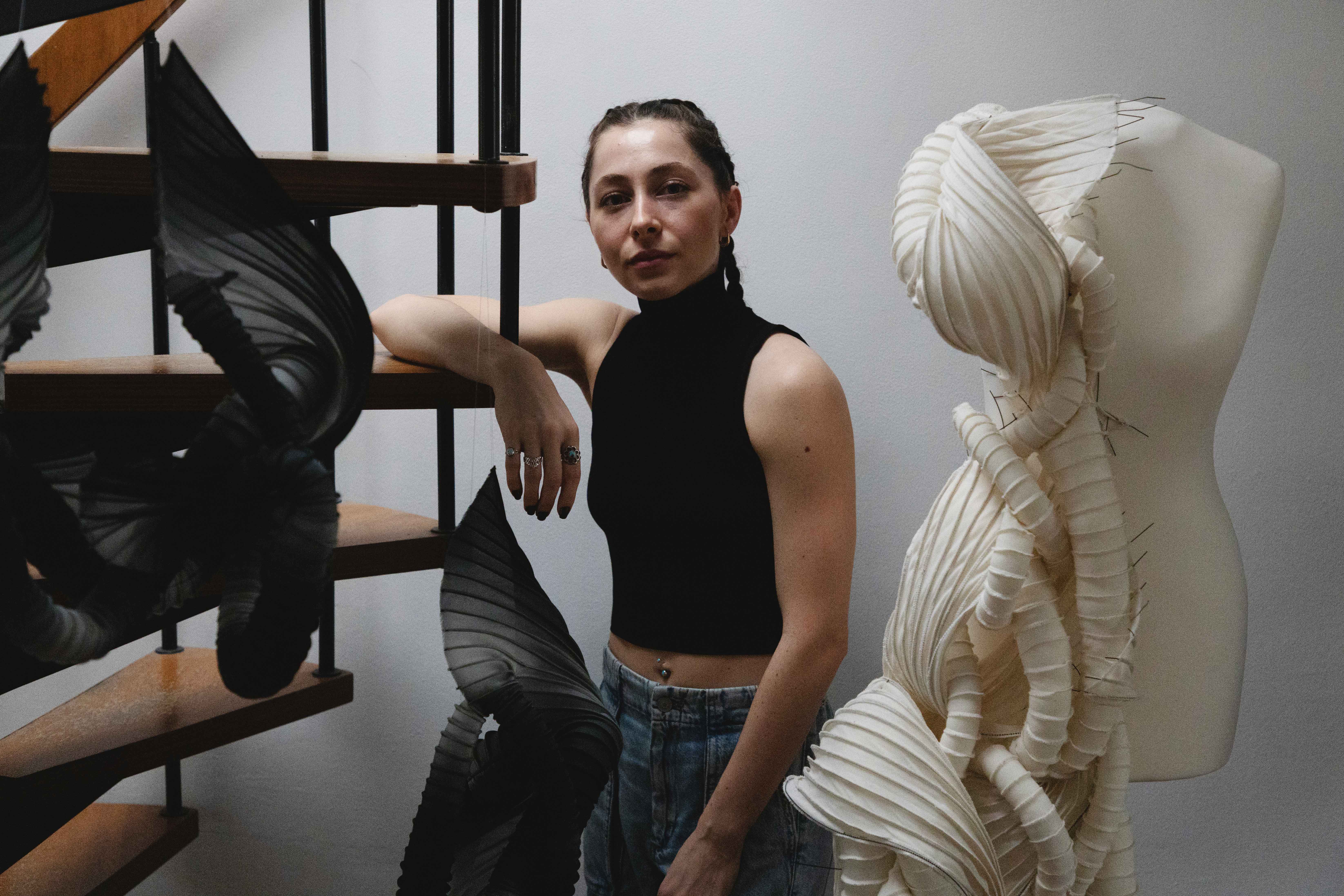 Kelsey is leaning against a circular staircase in her studio. She has a number of pieces of her work around her.  