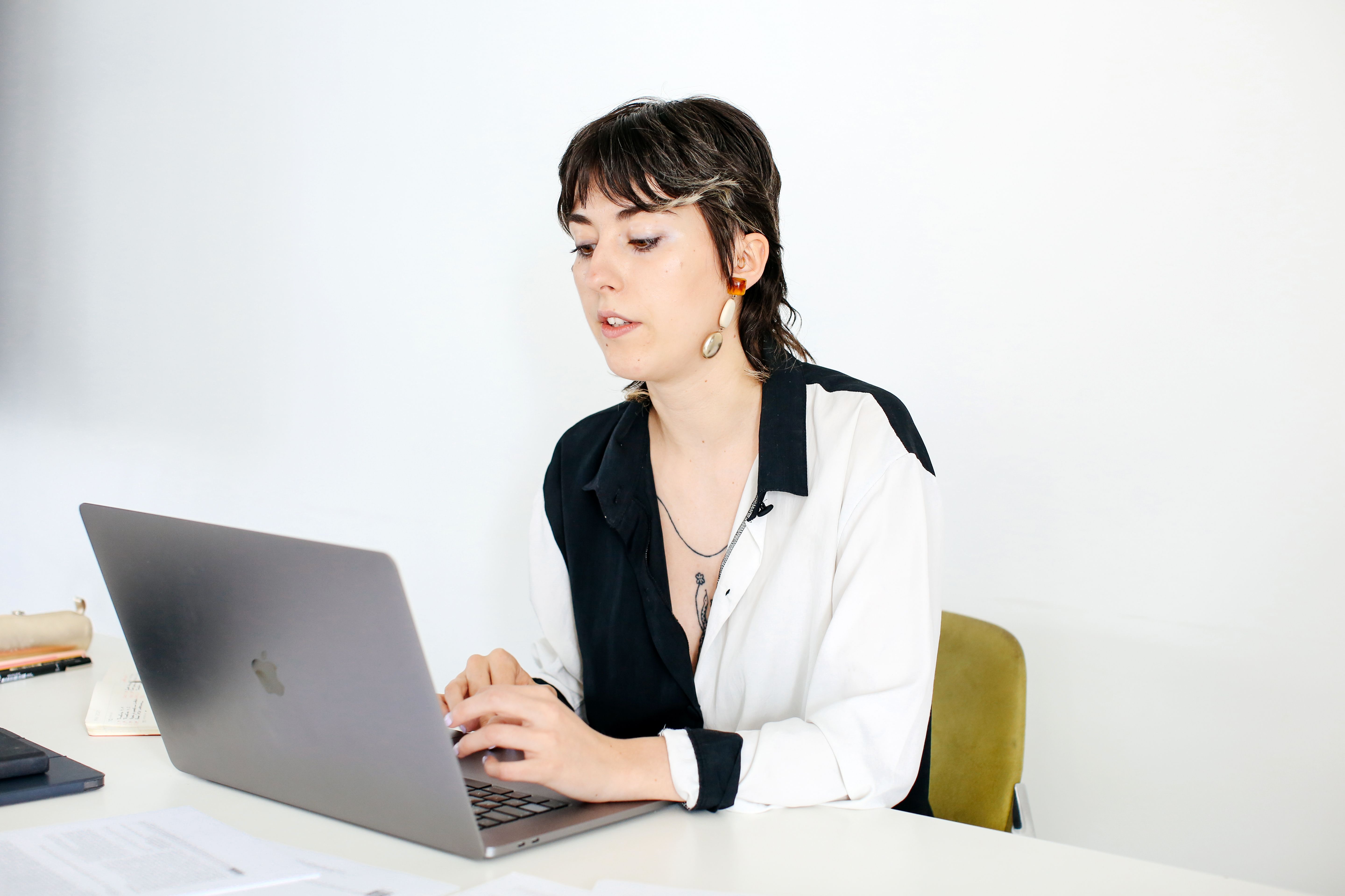 A woman with short dark hair and statement earrings is sitting at a desk, focused on her laptop. She is wearing a black and white shirt and appears to be working or typing in a bright, minimalist space.