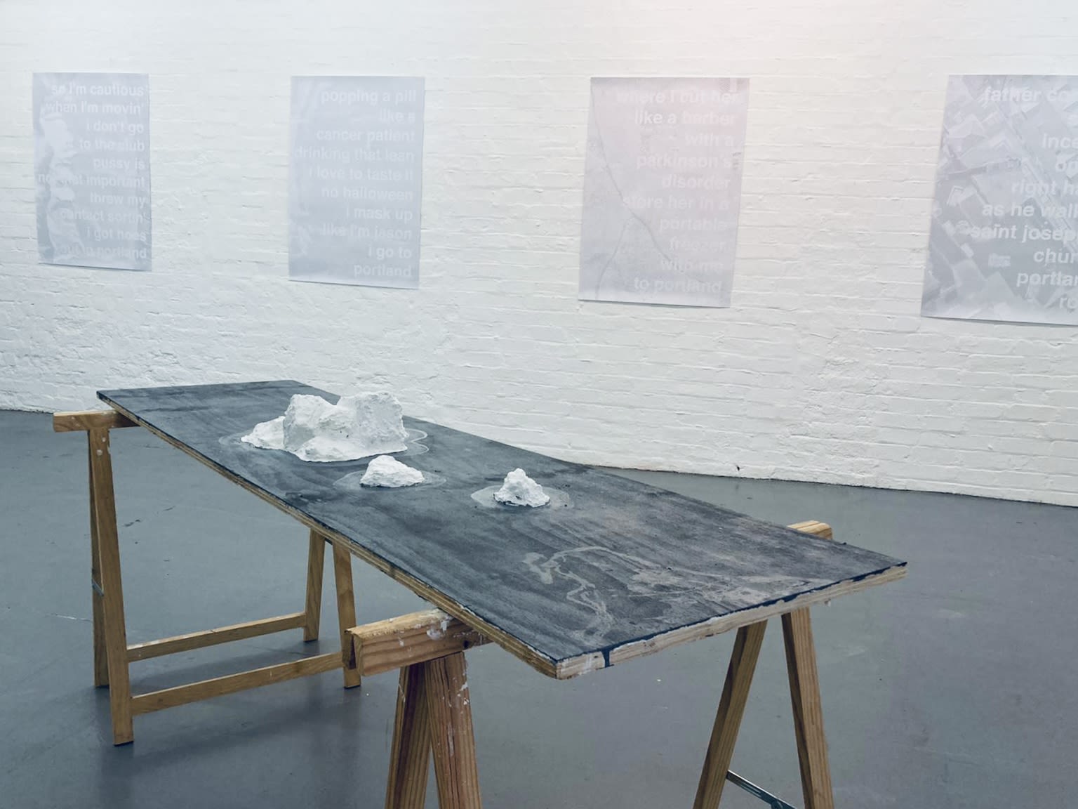 Black and wooden workbench with white sculptures on top in a room with a white brick wall covered in posters