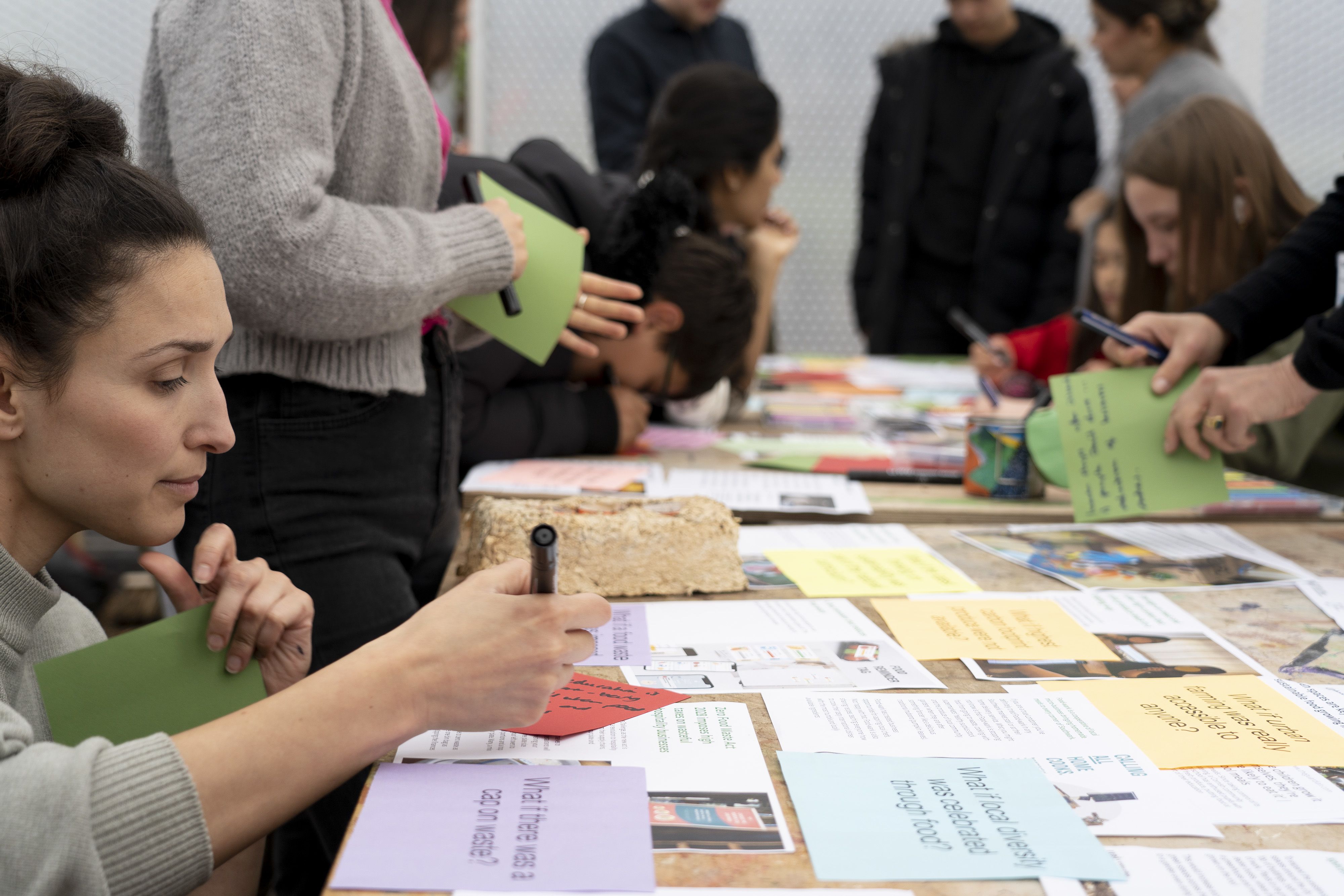 Close-up of a person kneeling to write something down on a piece of paper, while surrounded by others in a green house