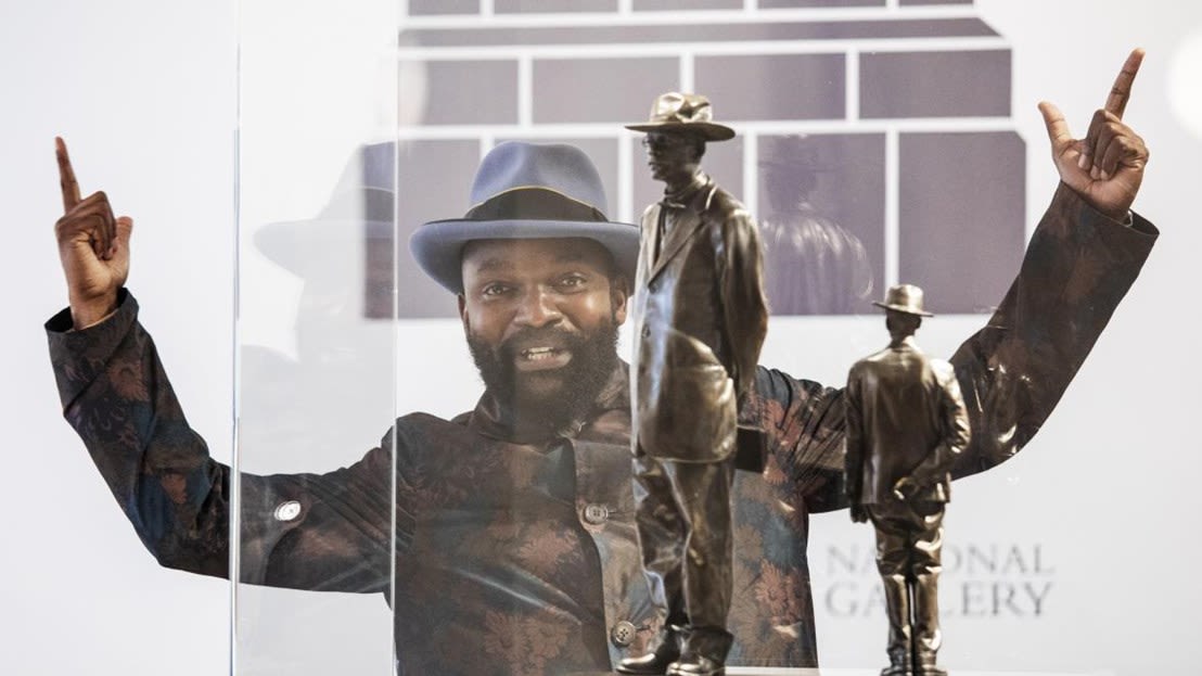 A Black man with his hands up with fingers pointing upwards in front of two bronze-coloured statues of men in hats.