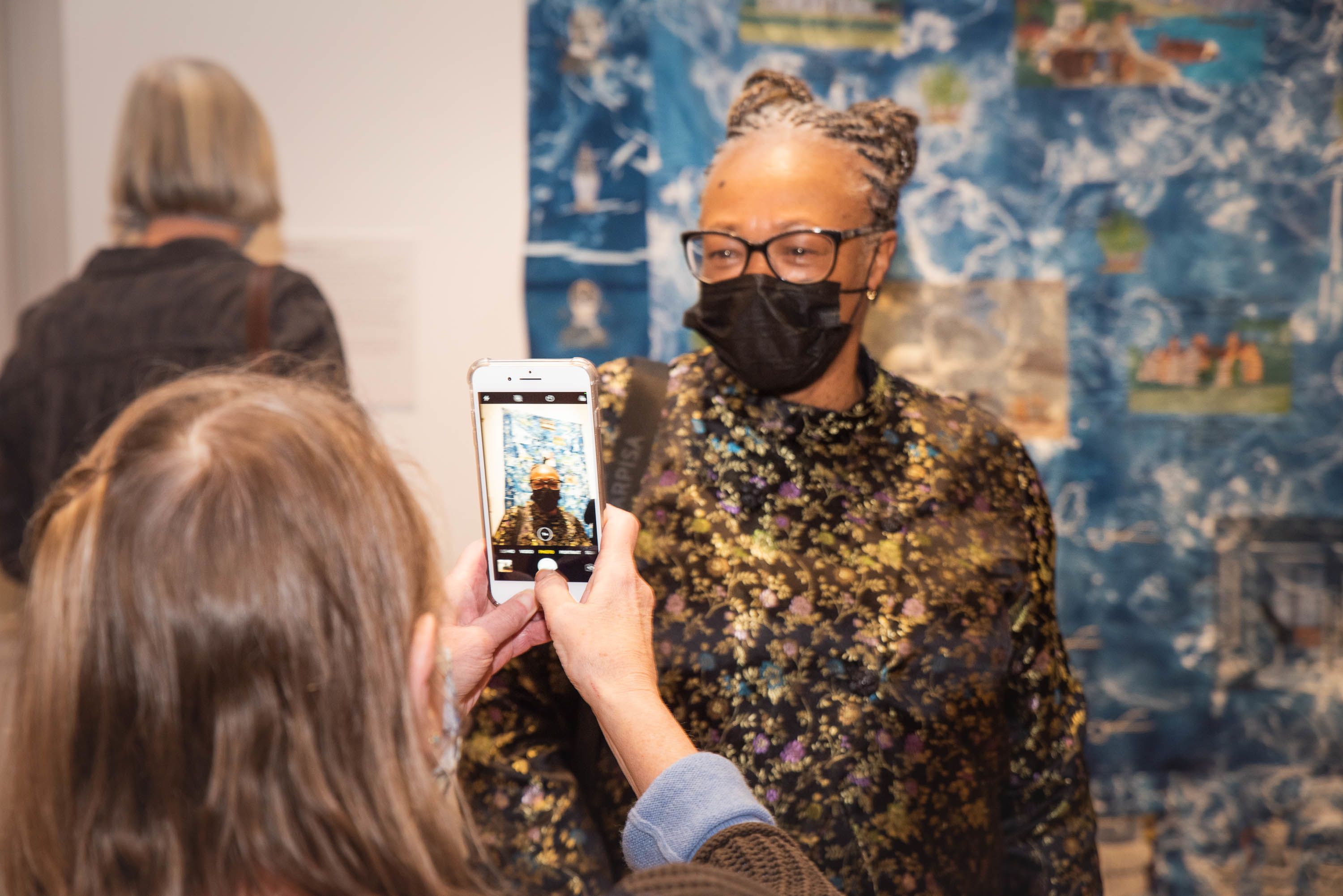 A person wearing glasses in front of an artwork with someone taking a photo of them on their phone