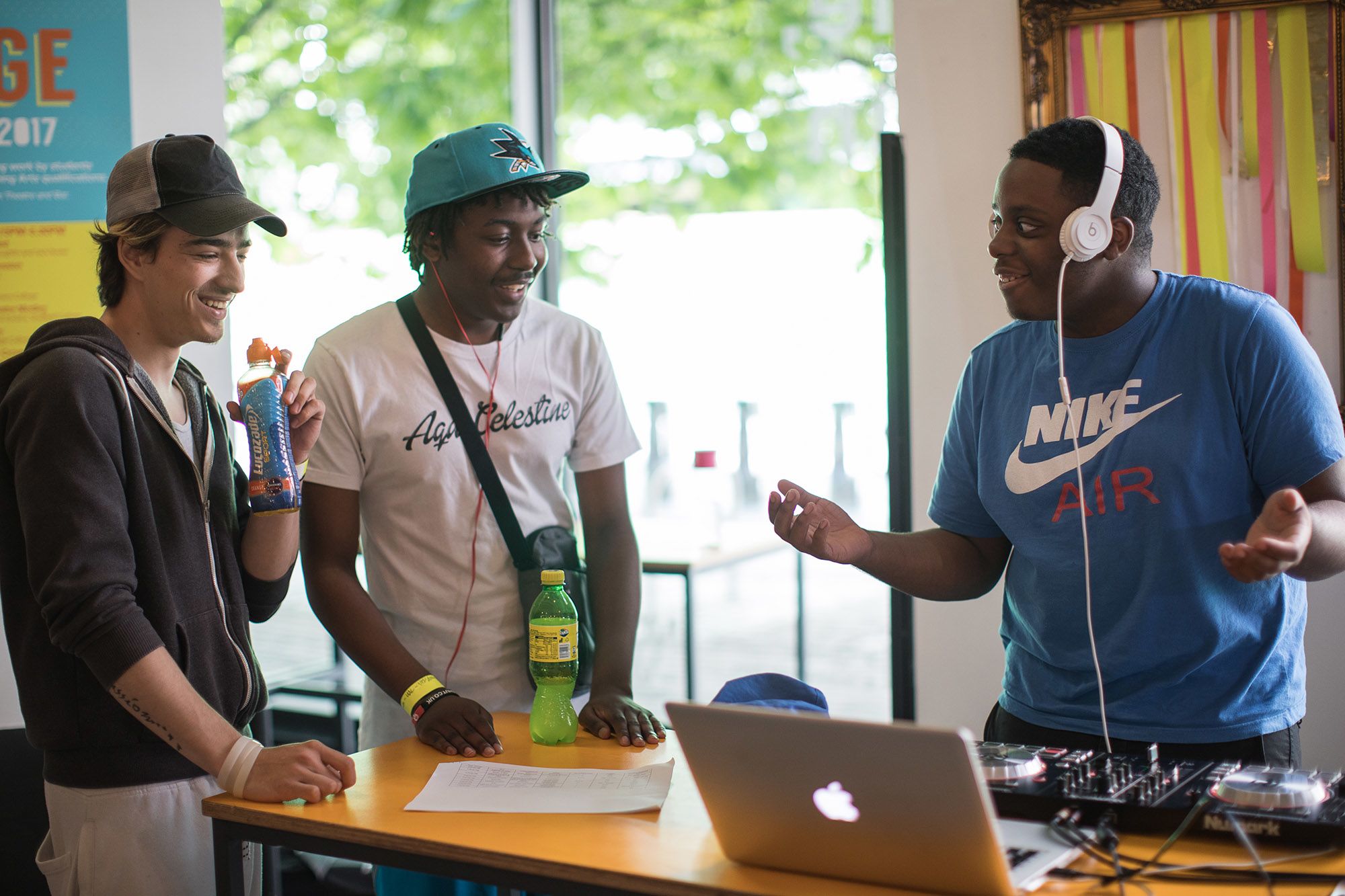 Three young men stand around a laptop and switch board wearing headphones and laughing