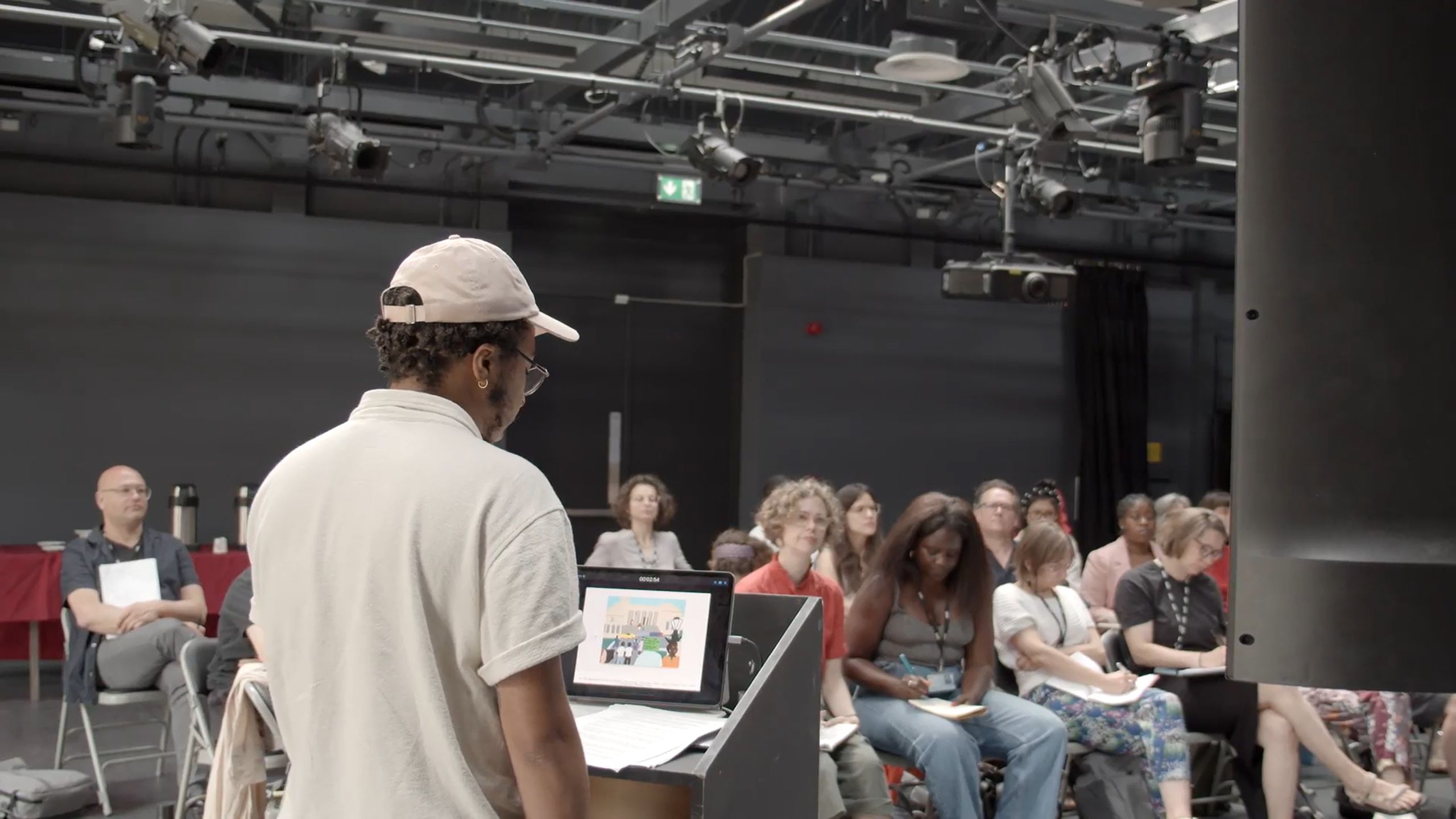 A man stands in front of a podium in front of a seated crowd giving a speech at Wimbledon College of Arts