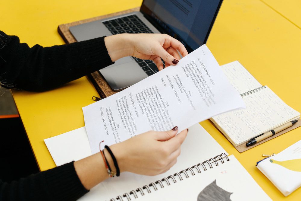 Close up of a woman's hands holding papers against a yellow desk with a laptop and notebooks