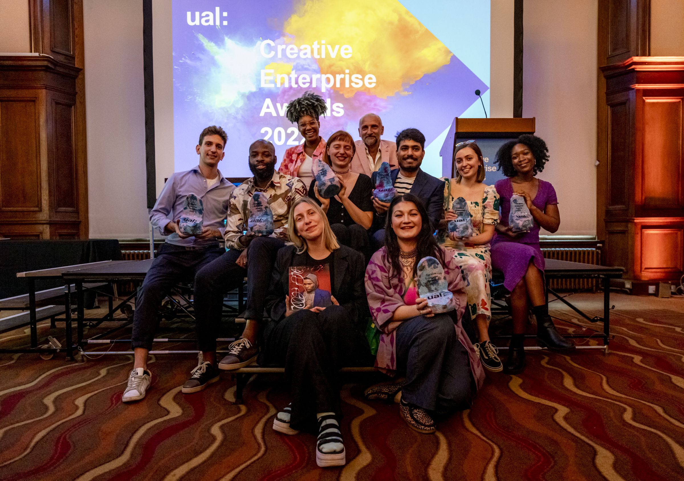 Winners of the Creative Enterprise Awards 2022 sit with Kemah Bob and Richard Sant, holding their trophies up and smiling for the camera