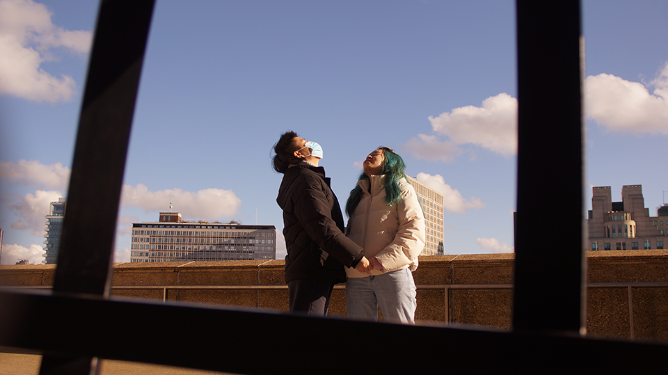 Video still showing two people wearing masks and holding hands while looking up