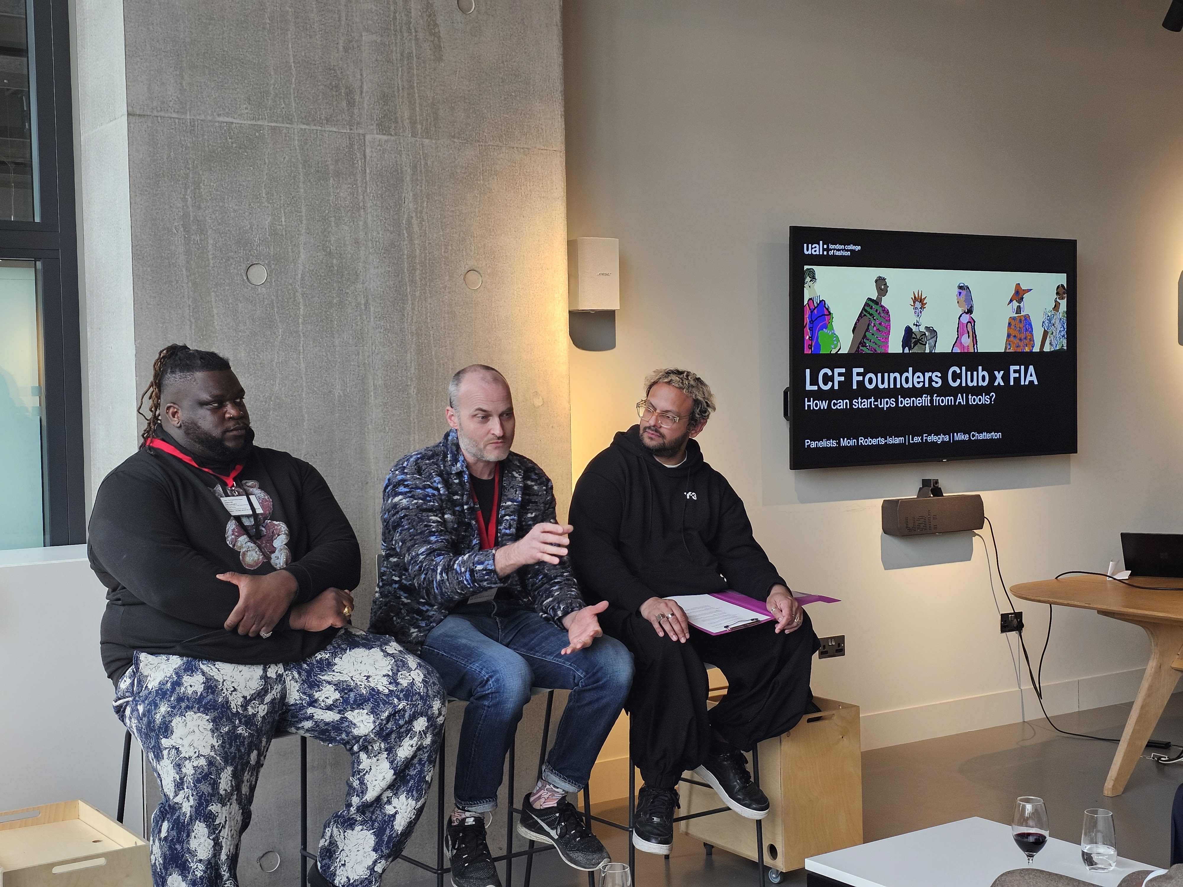 Three guest speakers talking and gesturing with their hands, sat on high stools next to a tv screen.