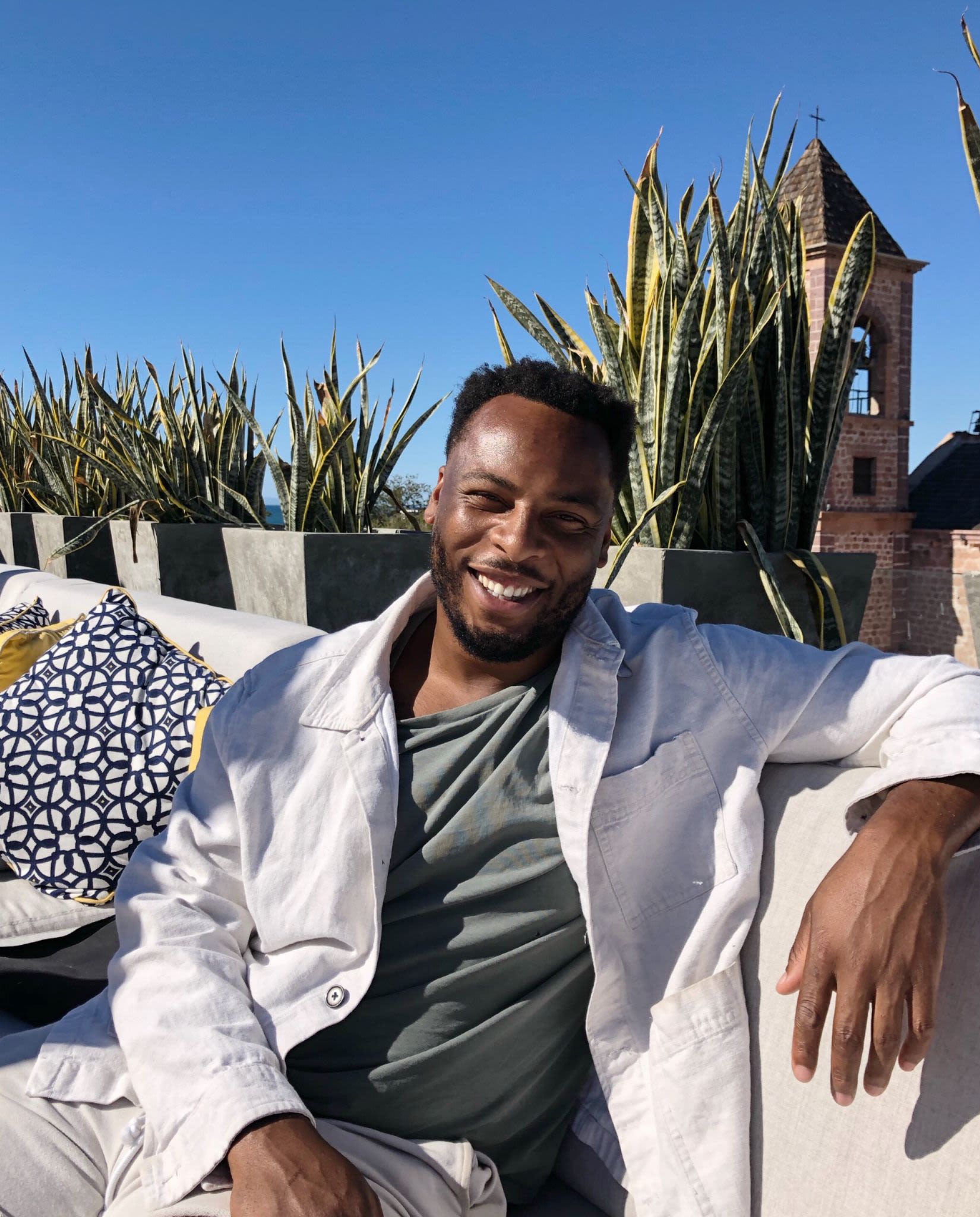 Photo of Solomon sitting outside in the sunshine smiling. Behind him are pots of plants along the top of the wall 