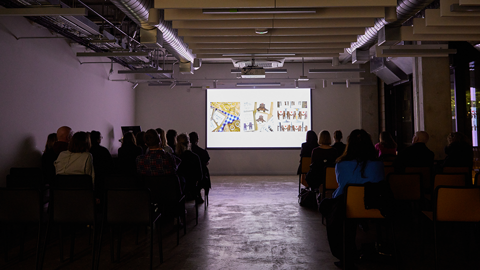 Long audience shot of people on the left and right and a big projector screen in the middle.