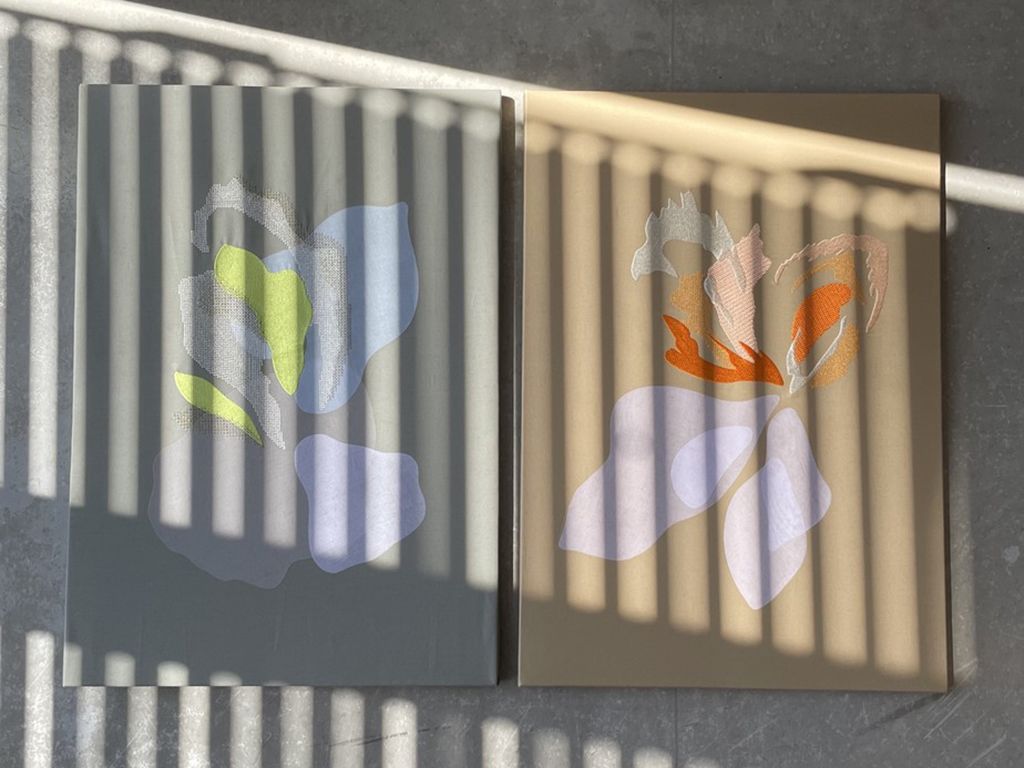 Two canvases hang on a grey wall. They are both displaying embroidered colourful flowers and are bathed in dappled light.